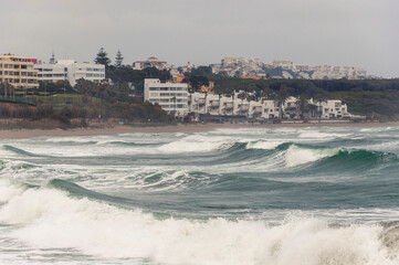 View of a spanish coastal resort town during a storm, with large turquoise waves in the foreground in cloudy weather