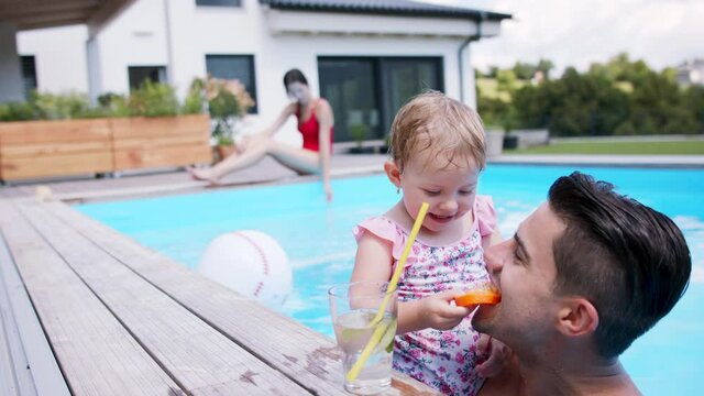 Small girl with father drinking lemonade in swimming pool outdoors in backyard garden.