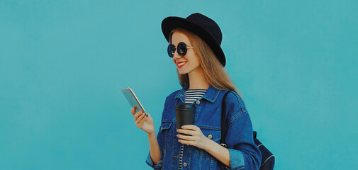 Fototapeta premium Portrait close up of smiling young woman with phone wearing a black round hat, denim jacket on a blue background