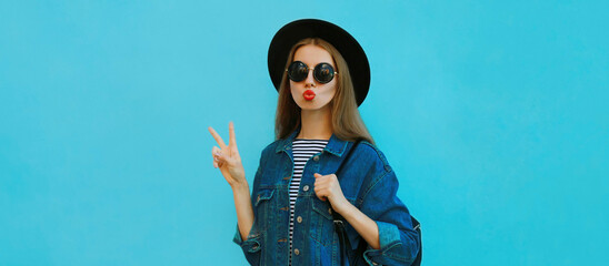 Portrait close up of young woman blowing her red lips sending sweet air kiss wearing a black round hat, denim jacket on a blue background