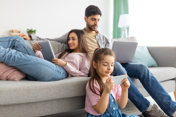 Gadgets addiction. Eastern family using different electronic devices, resting together in living room at home - Powered by Adobe