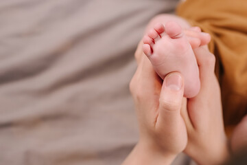 Close up of mother hand massaging foot newborn baby lying on comfortable grey bed at home. Caucasian loving mom takes care of her infant daughter. Copy space. Family, motherhood and child care concept