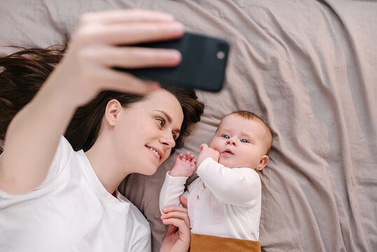 Happy Young Mother And Cute Sweet Baby Girl Having Video Chat Using Smartphone Mom Lying On Comfy Bed Near Toddler Enjoying Mobile Technology Sharing Motherhood Lifestyle With Friend On Social Media