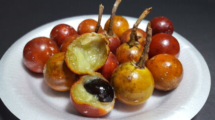 Tropical fruit growing in Trinidad and Tobago called Balata. A bunch is placed on a white plate on a black surface. One of the fruit is open to show the inside of the small brown, orange-like fruit. 