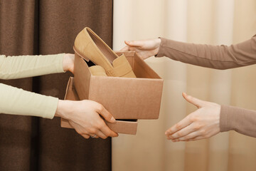 A cardboard box with suede loafers, which the hands of one girl pass to another against a draped...