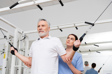 trainer supporting mature man training on pull rope exercising machine