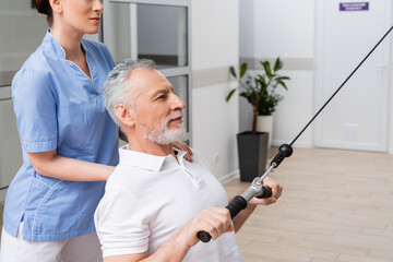 trainer assisting mature man training on pull cable exercising machine