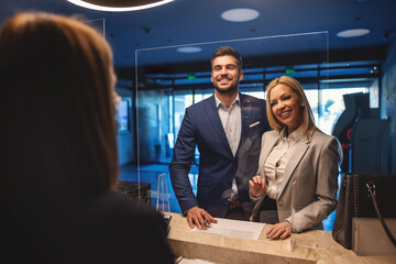 A happy adult business couple standing at a reception at the hotel and talking to the receptionist....