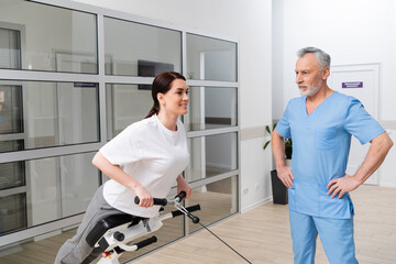 middle aged rehabilitologist standing with hands on hips near smiling woman training on exercising machine