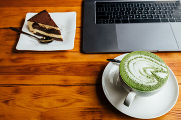 Top view of matcha coffee cup with piece of cake and laptop on brown textured table
