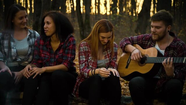 Multiethnic people wearing checked shirts sitting by campfire, one man playing guitar, women singing. Pan shot happy friends having fun in autumn forest at sunset. Concept of camping