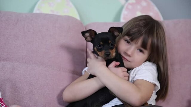 Adorable little girl hug her pet at home on sofa. Kid preparing for Easter at home during quarantine. Cute dog