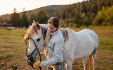 Young woman in warm jacket putting halter on white Arabian horse, blurred meadow and forest background
