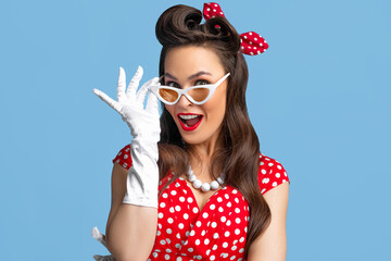 Portrait of excited pin up woman in polka dot dress, headband and gloves touching her sunglasses on...