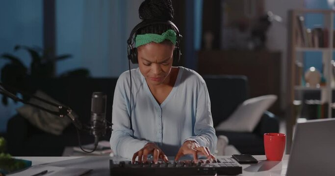Talented energetic young female musician wearing headphones creating new audiotracks on keyboard performing at home smiling for camera.