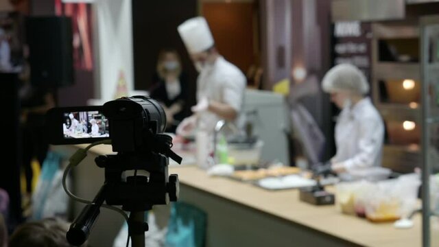 A video camera on a tripod is filming a culinary master class. Defocused videos of chefs in the kitchen, people out of focus. Blurred background with people.
