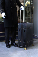 A uniformed doorman stands with a suitcase in his hand at the entrance to the hotel. The holiday...