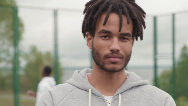 Close up tracking portrait shot of mixed race man with dreadlocks looking at camera on outdoor basketball court. People shooting hoops in background