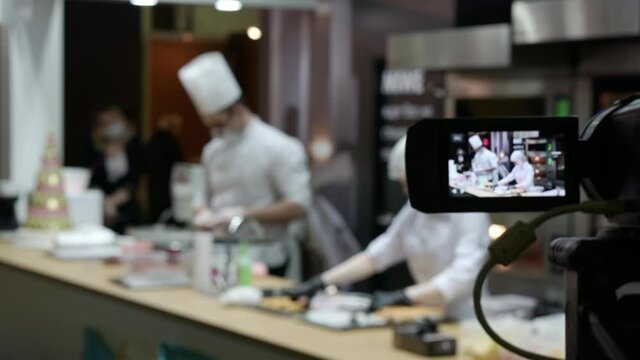 A video camera on a tripod is filming a culinary master class. Defocused videos of chefs in the kitchen, people out of focus. Blurred background with people.