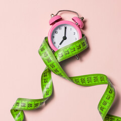 Pink alarm clock with green measuring tape, top view, copy space. Healthy lifestyle, weight loss.