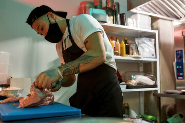 Focused young male cook wearing protective mask and gloves slicing raw pork meat on a plastic board while working in commercial kitchen