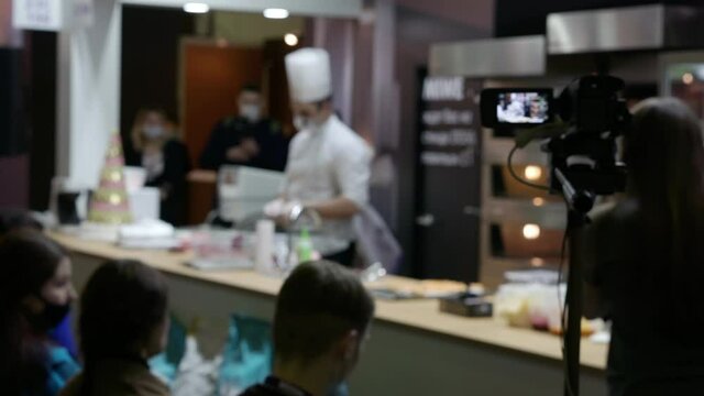 A video camera on a tripod is filming a culinary master class. Defocused videos of chefs in the kitchen, people out of focus. Blurred background with people.