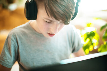 teenager in braces on teeth programs sitting at laptop on floor. boy in fashionable interior near trendy green plants from winter garden teaches lessons online at home.