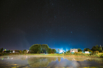 Fototapeta na wymiar Evaporation Over River Lake Near Houses In Village. Milky Way Galaxy In Night Starry Sky Above Lake River Landscape At Night.