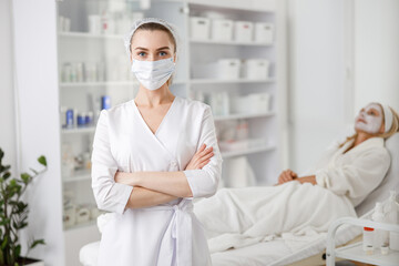 Female beauty doctor stands wears face mask while patience laying on background with cream or mask, in cosmetology clinic