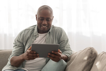 Mature black man spending time with digital tablet at home, browsing internet while relaxing on comfy couch
