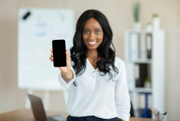 Cheerful black businesswoman holding smartphone with blank screen at office, mockup for mobile app or website design