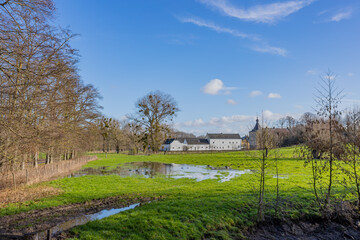 Fototapeta na wymiar Dutch farmland with a puddle of water on green grass, bare trees with Genhoes castle in the background, sunny day with a blue sky in Valkenburg aan de Geul in South Limburg, the Netherlands
