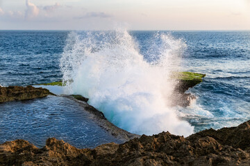 Wave breaking against cliff at Devil's Tear, on the coast of Nusa Lembongan, Bali, Indonesia. Pool of water on top of cliff in foreground. Blue ocean in the background. Sky and clouds in the distance.