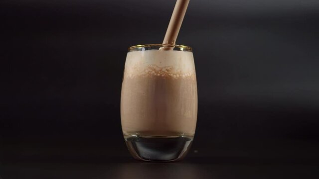 Drinking chocolate milk shake from straw. Close up on luxury glass filled with chocolate milk standing on dark background.