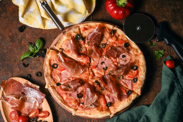 delicious round italian pizza with meat, olives, tomatoes, bell peppers and fresh herbs on a wooden round board on a brown background decorated with ingredients on the sides