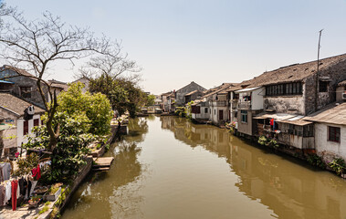 Fototapeta na wymiar Tongli, China - May 2, 2010: Brown water in canal with generic architecture white houses on both sides under light blue sky. Some green foliage and extra colors by drying laundry added.