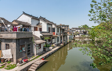 Fototapeta na wymiar Tongli, China - May 2, 2010: Greenish water in canal with generic architecture white houses on one side under light blue sky. Some green foliage and extra colors by drying laundry added.