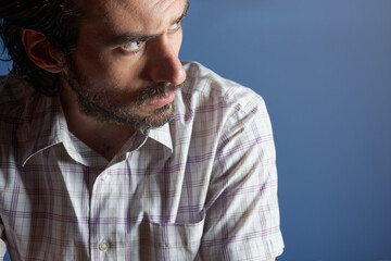 A young handsome serious bearded man with striped shirt sitting and looking away against light blue...