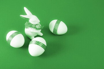 Easter Concept. Porcelain rabbit with three white eggs decorated with green ribbon on green paper background. Copy space.