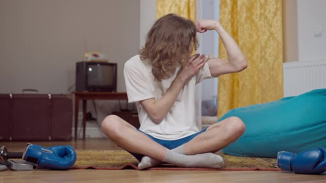 Portrait of positive young Caucasian man examining biceps sitting in retro home. Motivated confident guy satisfied with training result sitting on vintage carpet indoors. Sport and lifestyle in 80s.