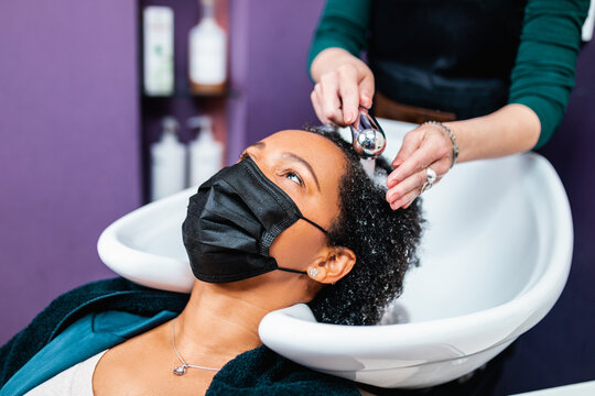 Professional hairdresser washing hair of a middle aged African American woman with protective face mask in hair salon.
