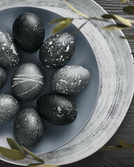 Black painted eggs for Easter on a blue plate, similar to stone, on a gray wooden background with dried flowers