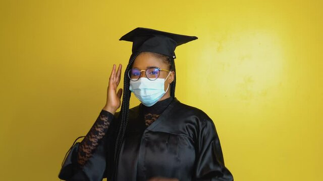The graduate student in a protective mask expresses with gestures the need to comply with quarantine security measures. Student in a black robe and hat on a yellow solid background looks at the camera