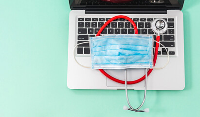 Surgical mask over a stethoscope placed above a computer keyboard on a pastel green background with good copyspace. Concept of technology and medicine with safety and prevention.