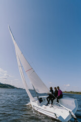 A beautiful white racing single-masted yacht is sailing against a beautiful river landscape with a blue sky. A man and two girls are on board.