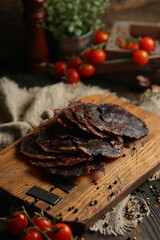 Delicious smoked sausage on a dark wooden board with tomatoes and spices. Delicious handmade snack made from natural ingredients. Pieces of dry pork, beef, chicken snacks. Meat products. Close-up.