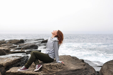 young woman in hoodie, jeans and simple sneakers sits on rocks near the stormy oceans with waves. local tourism, solo travel. mental health communication with nature.