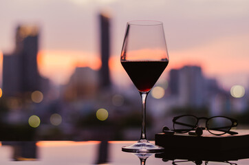 A glass of red wine with spectacles and ear pod on book put on table with city background. Stay home concept.