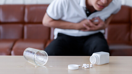 Close up shot of white plastic bottle of medicine, pills and glass of water fall on light brown wooden table when old man hold left chest with two hands has heart attack failure in blurred background