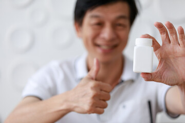 Close up shot of red pills vitamin food supplement in small glass bottle with wood cap was hold by Asian old middle aged male wears white shirt shows thumb up smiling happily in blurred background